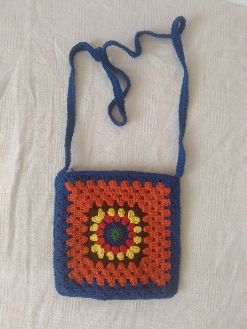 May include: A crocheted crossbody bag with a blue strap and a colorful granny square design. The square features a circular pattern in shades of orange, yellow, green, brown, and red.