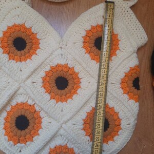May include: Cream-colored crochet tote bag featuring sunflower motifs in orange, brown, and black. The bag is approximately 28 inches long, with a strap for carrying. The bag is handmade and has a unique, textured appearance.