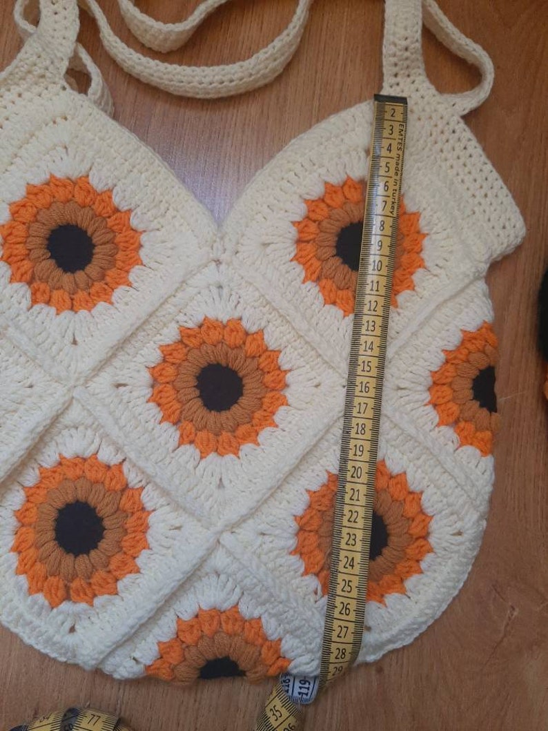 May include: Cream-colored crochet tote bag featuring sunflower motifs in orange, brown, and black. The bag is approximately 28 inches long, with a strap for carrying. The bag is handmade and has a unique, textured appearance.