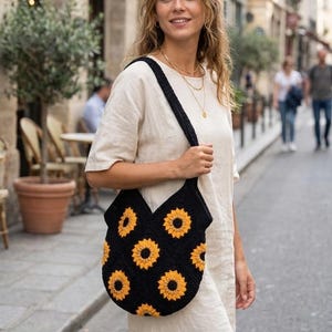 May include: Black crochet tote bag with a sunflower pattern in yellow and orange. The bag has a wide strap and is carried over the shoulder. The bag is made of a soft, textured material.