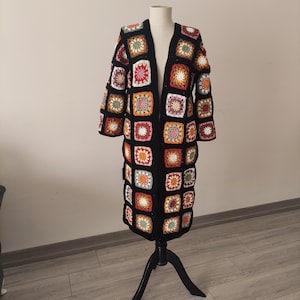 Puede incluir: Un cárdigan largo de crochet con frente abierto, exhibido en un maniquí. El cárdigan presenta una base negra con patrones cuadrados coloridos en tonos de rojo, naranja, amarillo, verde y blanco. La prenda se exhibe sobre un soporte negro.