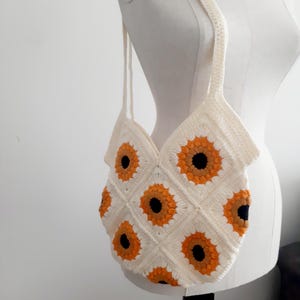 May include: Cream-colored crochet tote bag featuring a sunflower pattern. The bag has a rectangular shape with a diamond pattern of sunflowers in orange and black. The bag has two long straps for carrying.