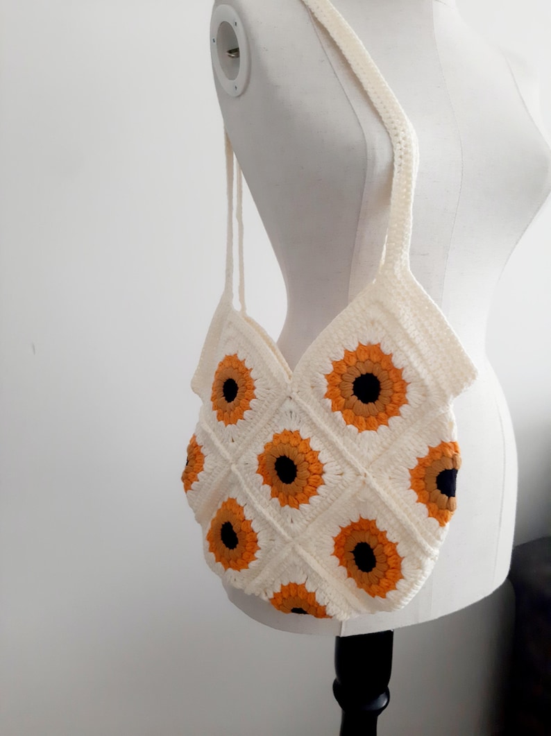 May include: Cream-colored crochet tote bag featuring a sunflower pattern. The bag has a rectangular shape with a diamond pattern of sunflowers in orange and black. The bag has two long straps for carrying.