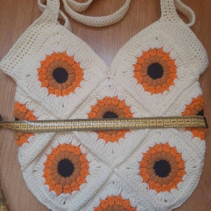 May include: Cream-colored crochet bag with a sunflower design. The bag features a repeating pattern of orange and brown sunflowers on a white background. The bag has a strap and measures approximately 11 inches wide.