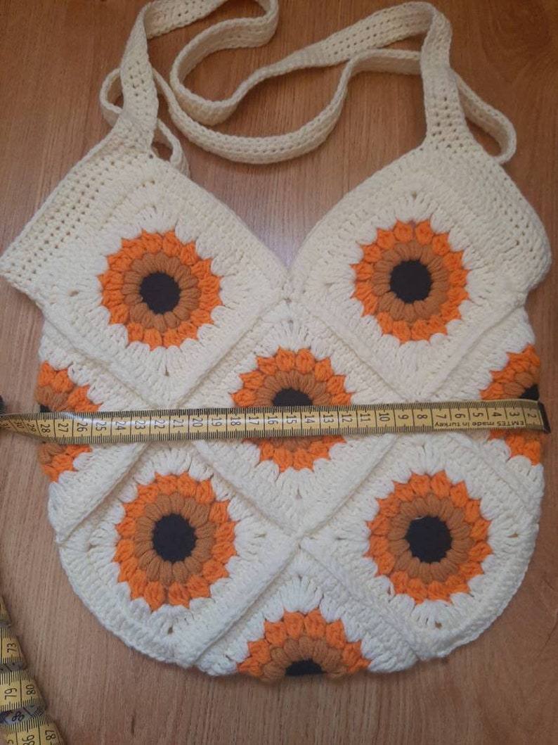 May include: Cream-colored crochet bag with a sunflower design. The bag features a repeating pattern of orange and brown sunflowers on a white background. The bag has a strap and measures approximately 11 inches wide.