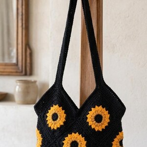 May include: Black crochet tote bag with yellow sunflower appliques. The bag is hanging on a wooden hook. The bag has two long straps and a V-shaped opening. The sunflowers are in full bloom.