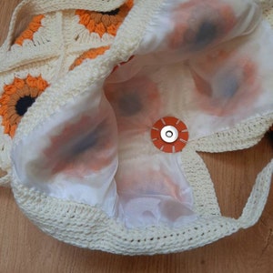 May include: Cream-colored crochet bag with sunflower motifs in orange, brown, and black. The bag features a white satin lining and a circular orange magnetic closure. The bag's strap is also crocheted.