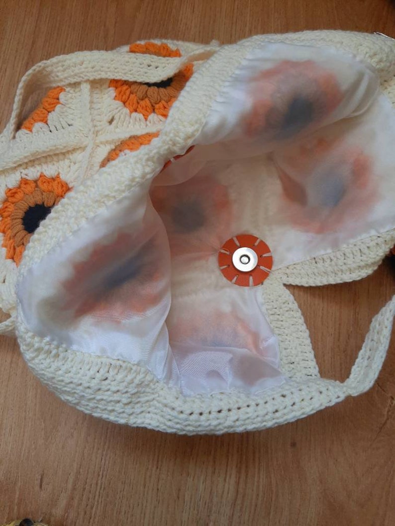 May include: Cream-colored crochet bag with sunflower motifs in orange, brown, and black. The bag features a white satin lining and a circular orange magnetic closure. The bag's strap is also crocheted.