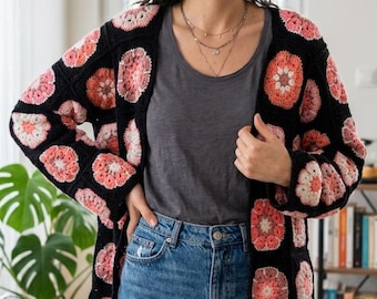 Könnte beinhalten: Schwarze Häkeljacke mit einem Blumenmuster in Rosa und Weiß. Der offene Pullover wird über einem grauen Oberteil und einer blauen Jeans getragen. Die Strickjacke ist ein einzigartiges, handgefertigtes Stück.