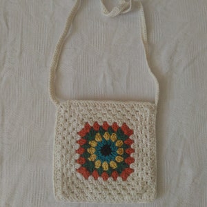 May include: A white crocheted bag with a colorful granny square design. The bag has a long strap for carrying over the shoulder.