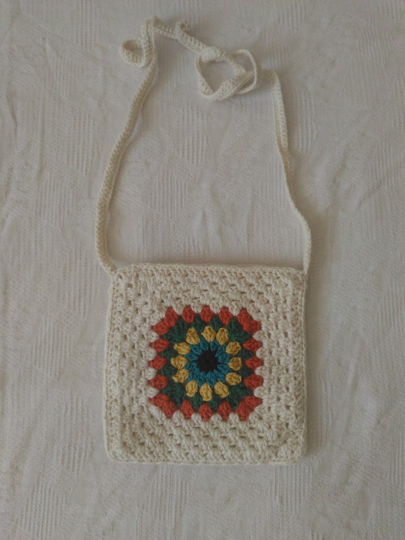 May include: A white crocheted bag with a colorful granny square design. The bag has a long strap for carrying over the shoulder.