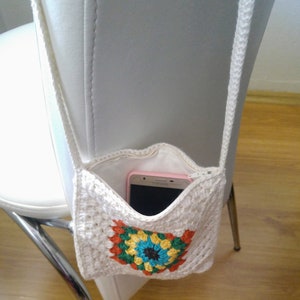 May include: A white crocheted crossbody bag with a colorful granny square design. The bag has a zipper closure and is holding a pink smartphone.