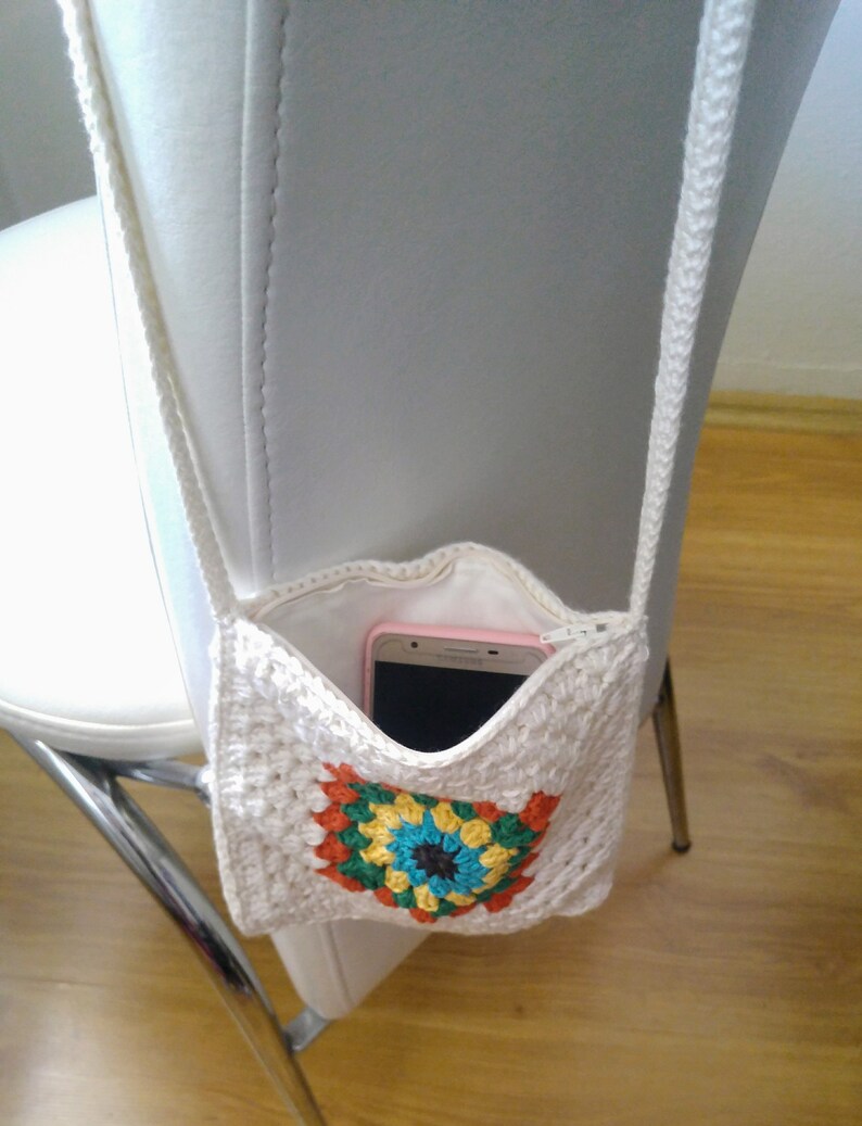May include: A white crocheted crossbody bag with a colorful granny square design. The bag has a zipper closure and is holding a pink smartphone.