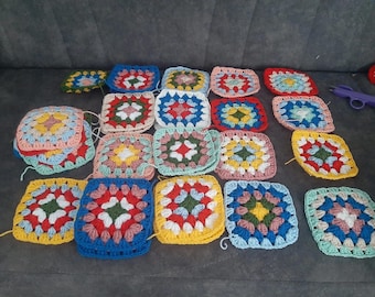 May include: A selection of colourful, crocheted granny squares in various stages of completion. The squares feature a variety of colours, including red, blue, yellow, and green, and are arranged on a grey surface. A pair of purple scissors is also visible.