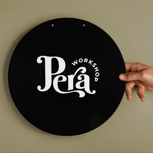 May include: A black circular sign with white text that reads "Pera Workshop". The sign is hanging on a light green wall.