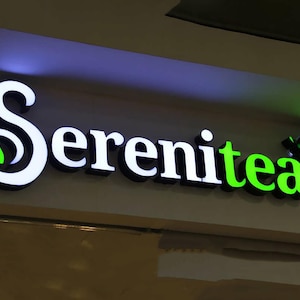 以下が含まれることがあります： 「Serenitea」というお茶屋さんの電飾看板。 白い3D文字に黒い縁取りが施されています。「Serenitea」の文字は、スタイリッシュなフォントで、"tea"の部分は明るい緑色です。 緑色の葉のグラフィックがデザインに組み込まれています。