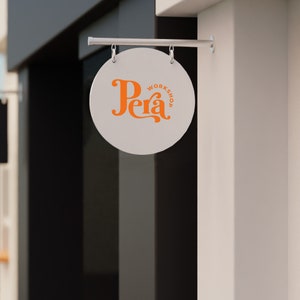 May include: A white round sign with a silver metal frame hanging from a ceiling. The sign says "Pera Workshop" in orange lettering.