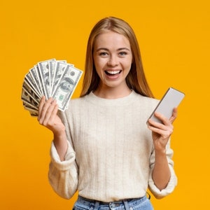 Puede incluir: Una mujer con un suéter blanco sostiene un abanico de billetes de dólar estadounidense en una mano y un teléfono inteligente en la otra. Ella sonríe y mira a la cámara. El fondo es amarillo brillante.
