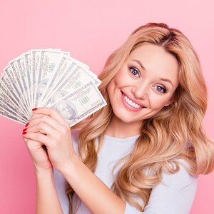 May include: A woman with long blonde hair smiles while holding a fan of US dollar bills in front of her face. The background is a solid pink.