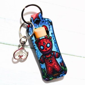 May include: A blue and red keychain with a sugar skull design holds a lip balm. The keychain has a silver heart-shaped charm.