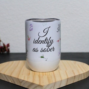 May include: White stainless steel tumbler with a black inscription that reads "I identify as sober". The tumbler is decorated with colorful cocktail and drink icons.