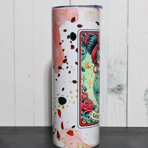 Puede incluir: Un vaso de acero inoxidable blanco con un dise&ntilde;o de acuarela rosa y dorado. El vaso presenta un gr&aacute;fico de una mujer con una cara de calavera de az&uacute;car y el texto "La Calavera".