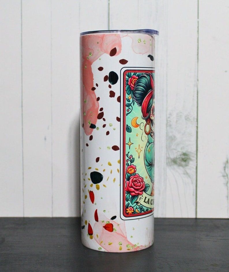 Puede incluir: Un vaso de acero inoxidable blanco con un dise&ntilde;o de acuarela rosa y dorado. El vaso presenta un gr&aacute;fico de una mujer con una cara de calavera de az&uacute;car y el texto "La Calavera".