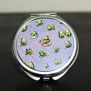 May include: A silver compact mirror with a purple background and a pattern of cartoon avocados in various poses. The avocados are wearing sunglasses, swimming, and relaxing on a flamingo float.