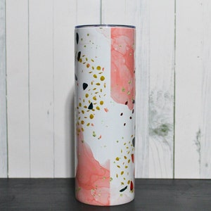 Puede incluir: Un vaso de acero inoxidable blanco y rosa con un dise&ntilde;o abstracto dorado y negro.