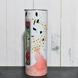 Puede incluir: Un vaso blanco con un dise&ntilde;o rosa y dorado. El dise&ntilde;o presenta un patr&oacute;n floral y purpurina dorada. El vaso tiene una tapa transparente.