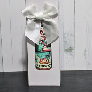 Puede incluir: Caja de regalo blanca con un lazo de sat&eacute;n blanco. La caja contiene una botella con una ilustraci&oacute;n colorida de un esqueleto con una camisa azul y el texto "LA CHINGON".