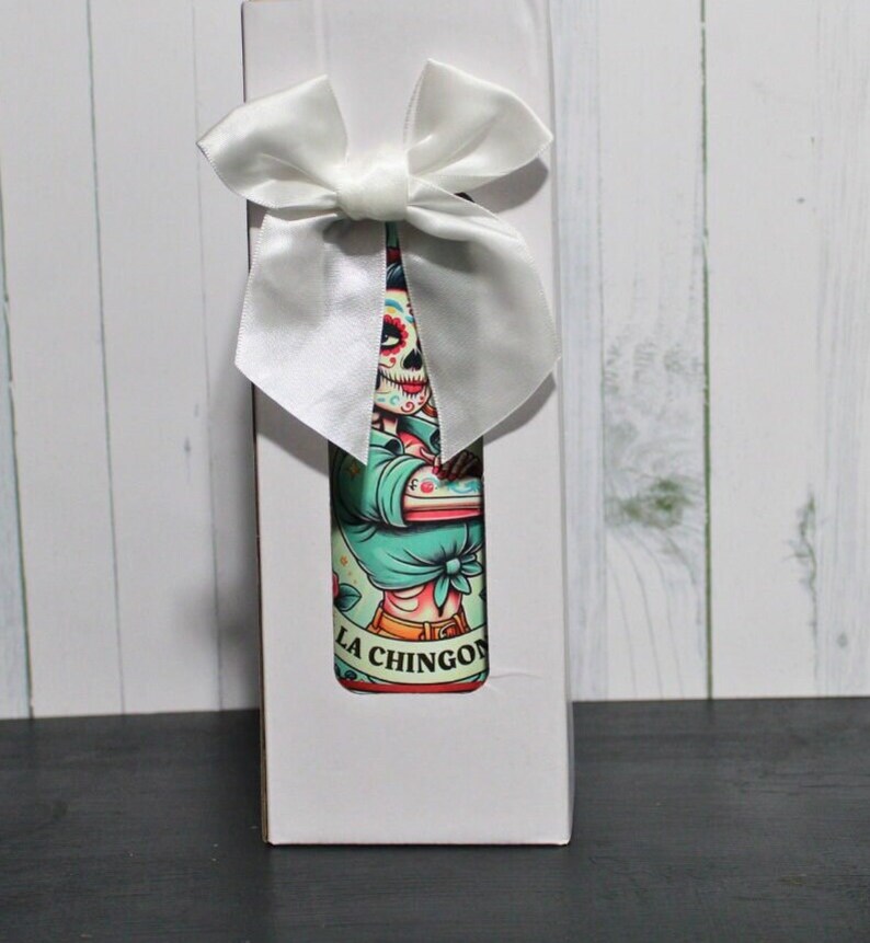 Puede incluir: Caja de regalo blanca con un lazo de sat&eacute;n blanco. La caja contiene una botella con una ilustraci&oacute;n colorida de un esqueleto con una camisa azul y el texto "LA CHINGON".