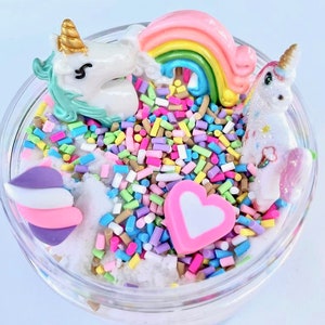 K&ouml;nnte beinhalten: Ein transparentes Glas gef&uuml;llt mit wei&szlig;em, fluffigem Schleim, Regenbogenstreuseln und Einhornfiguren. Der Schleim ist mit einem rosa Herz, einem Regenbogen und einem Einhorn mit goldenem Horn verziert.