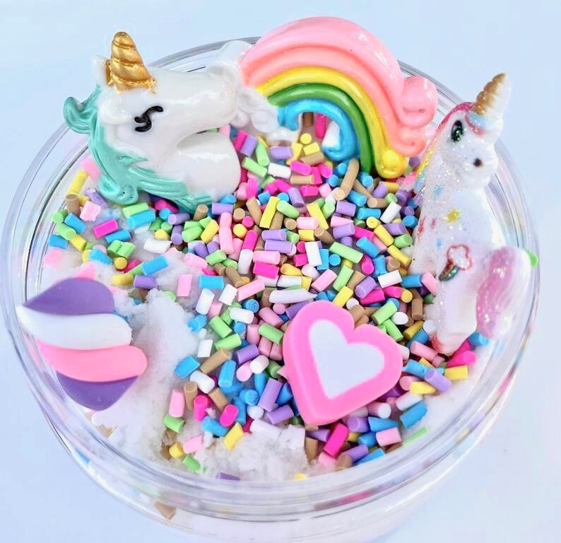 K&ouml;nnte beinhalten: Ein transparentes Glas gef&uuml;llt mit wei&szlig;em, fluffigem Schleim, Regenbogenstreuseln und Einhornfiguren. Der Schleim ist mit einem rosa Herz, einem Regenbogen und einem Einhorn mit goldenem Horn verziert.