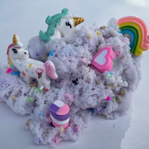 K&ouml;nnte beinhalten: Lilafarbener, flauschiger Schleim mit wei&szlig;en und regenbogenfarbenen Streuseln, einem rosa Herz, einem Regenbogen und zwei wei&szlig;en Einhorn-Figuren.