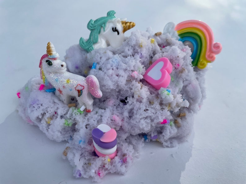K&ouml;nnte beinhalten: Lilafarbener, flauschiger Schleim mit wei&szlig;en und regenbogenfarbenen Streuseln, einem rosa Herz, einem Regenbogen und zwei wei&szlig;en Einhorn-Figuren.