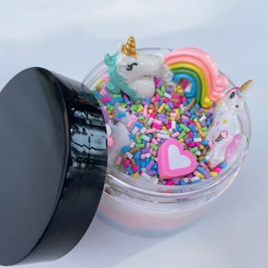 K&ouml;nnte beinhalten: Ein transparentes Glas gef&uuml;llt mit wei&szlig;em Schleim, bunten Streuseln, einem rosa Herz und zwei Einhorn-Figuren. Das Glas hat einen schwarzen Deckel.