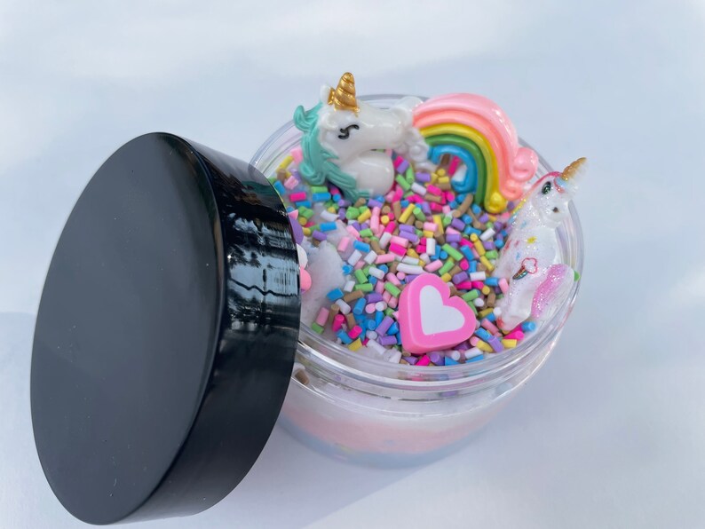 K&ouml;nnte beinhalten: Ein transparentes Glas gef&uuml;llt mit wei&szlig;em Schleim, bunten Streuseln, einem rosa Herz und zwei Einhorn-Figuren. Das Glas hat einen schwarzen Deckel.