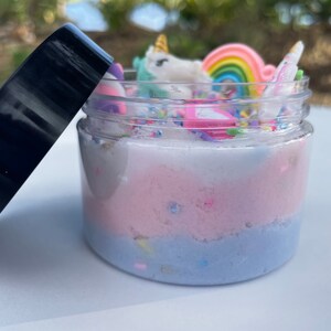K&ouml;nnte beinhalten: Ein transparentes Glas gef&uuml;llt mit mehrschichtigem, mehrfarbigem Schleim. Der Schleim ist wei&szlig;, rosa und blau. Das Glas ist mit einem schwarzen Deckel versehen und mit Einhorn- und Regenbogen-Anh&auml;ngern verziert.