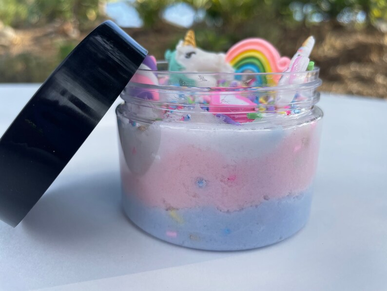 K&ouml;nnte beinhalten: Ein transparentes Glas gef&uuml;llt mit mehrschichtigem, mehrfarbigem Schleim. Der Schleim ist wei&szlig;, rosa und blau. Das Glas ist mit einem schwarzen Deckel versehen und mit Einhorn- und Regenbogen-Anh&auml;ngern verziert.