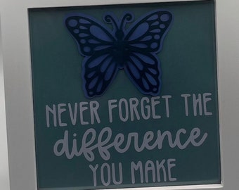 May include: A framed inspirational quote with a blue butterfly cutout. The quote reads "Never forget the difference you make."