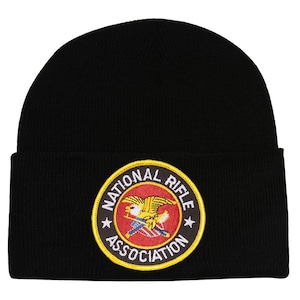 Op de afbeelding: Zwarte gebreide beanie met een rood, wit en blauw geborduurde patch met de tekst "National Rifle Association" in een cirkel met een gele rand.