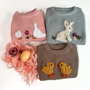 May include: Three knitted jumpers with embroidered animal designs. The pink jumper has a goose and a chick, the blue jumper has a rabbit, and the brown jumper has two chicks.
