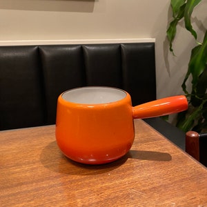 May include: An orange enameled saucepan with a single handle. The interior is white, and the exterior has a gradient from a darker orange at the base to a lighter orange at the rim and handle. The saucepan is sitting on a wooden table.