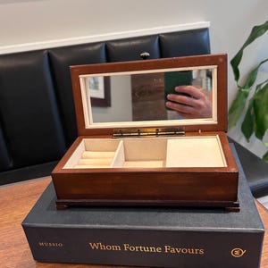 May include: A rectangular wooden jewelry box with a dark brown finish, featuring an interior mirror on the lid and a beige velvet-lined compartment with ring rolls. The box rests on a dark blue book titled "Mussio Whom Fortune Favours."