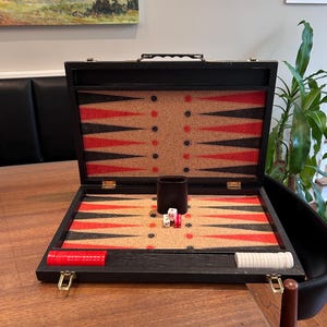 May include: A black backgammon board case with a cork-textured interior featuring red and black triangular points. The set includes a black dice cup, two white dice, one red die, and stacks of red and white checkers stored in the side compartments.
