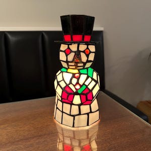 May include: A stained glass snowman lamp with a black top hat and red trim. The snowman's body is composed of white, red, and green glass pieces. The lamp is illuminated, casting a warm glow.
