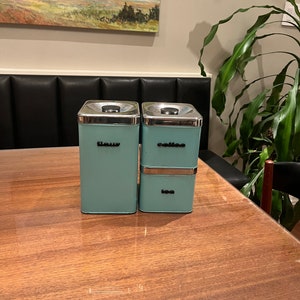 May include: Three aqua-colored metal canisters with silver lids. The canisters are labeled "flour", "coffee", and "tea" in a simple, black font. The canisters are stacked on a wooden table.