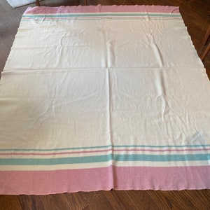 May include: A large, off-white blanket with a pink border and stripes. The blanket features a light blue and pink stripe design. The blanket is folded on a wooden floor.
