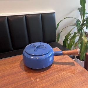 May include: A round, blue enameled metal pot with a matching lid featuring a cross-shaped handle. The pot has a single, straight, light brown wooden handle extending from the side, providing a contrast to the smooth, glossy blue finish of the body.